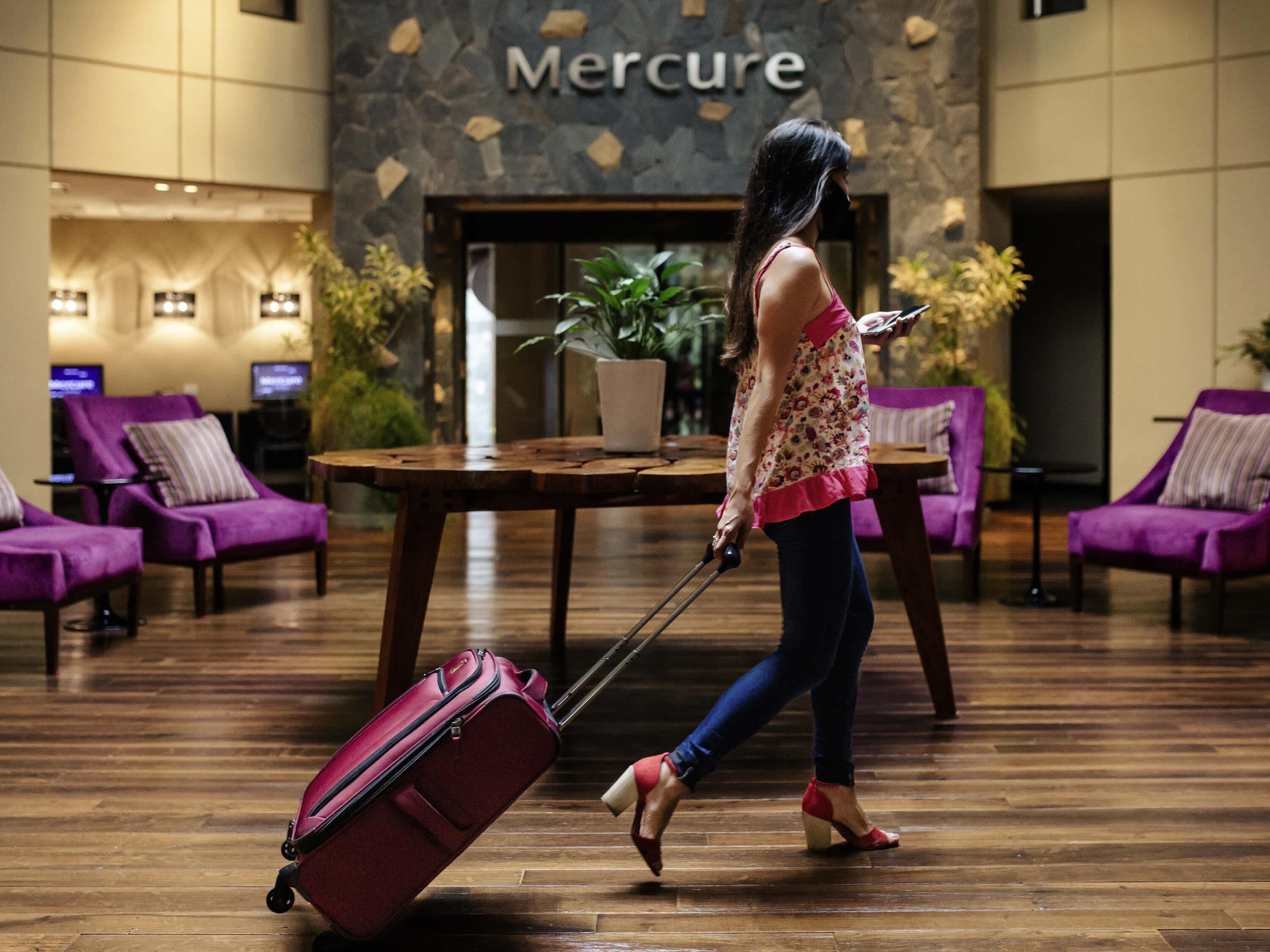 Une femme marchant dans un hall, une valise à la main, devant des fauteuils violets et un mur de pierre sur lequel est inscrit le mot « Mercure ».