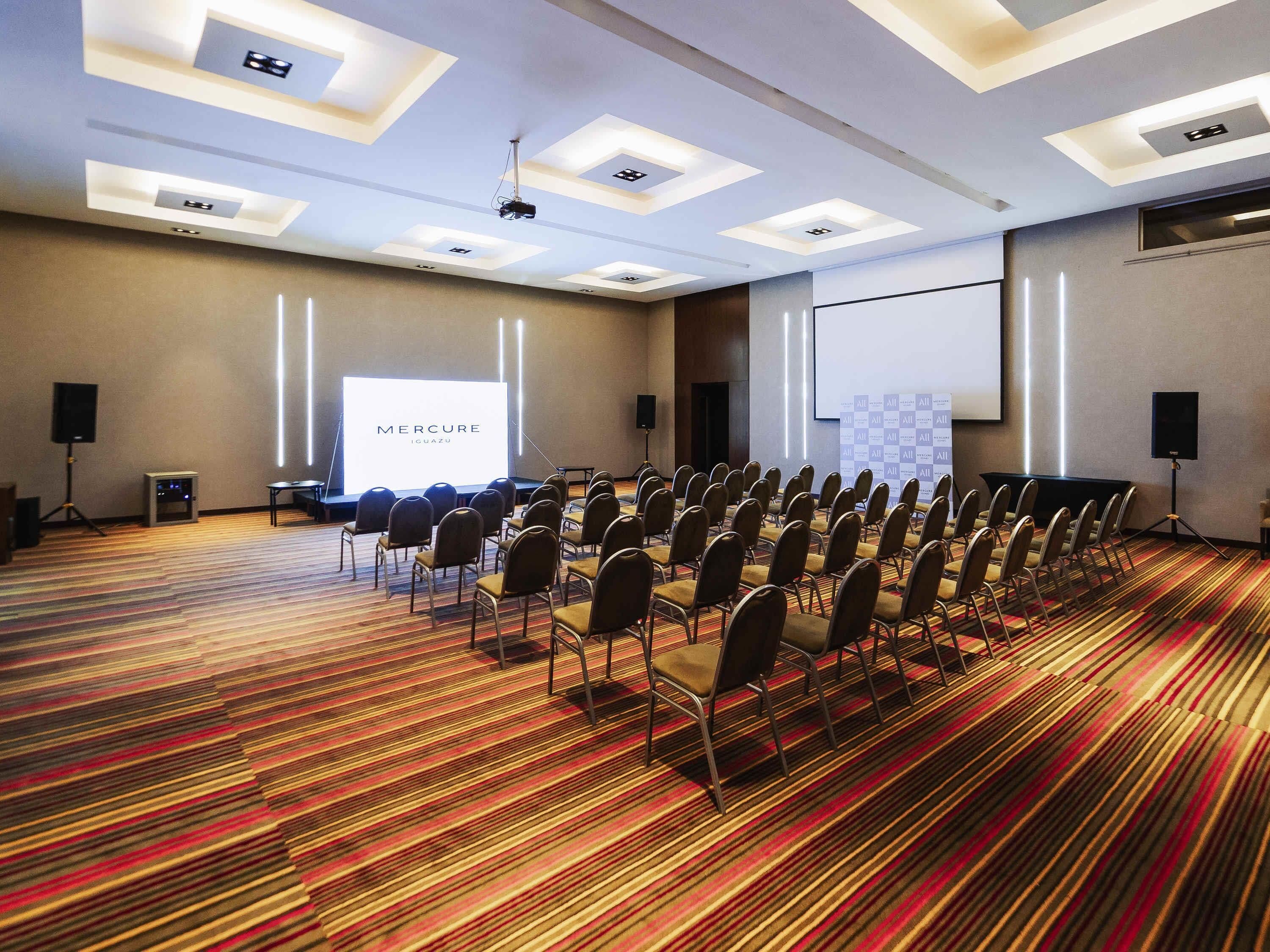 Un salón de conferencias con filas de sillas, una pantalla de proyector y una pantalla grande que muestra 'MERCURE IQUAZU'.
