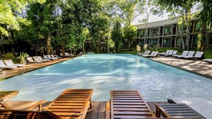 Una piscina con tumbonas y sillones de madera, rodeada de árboles y un edificio con grandes ventanales.