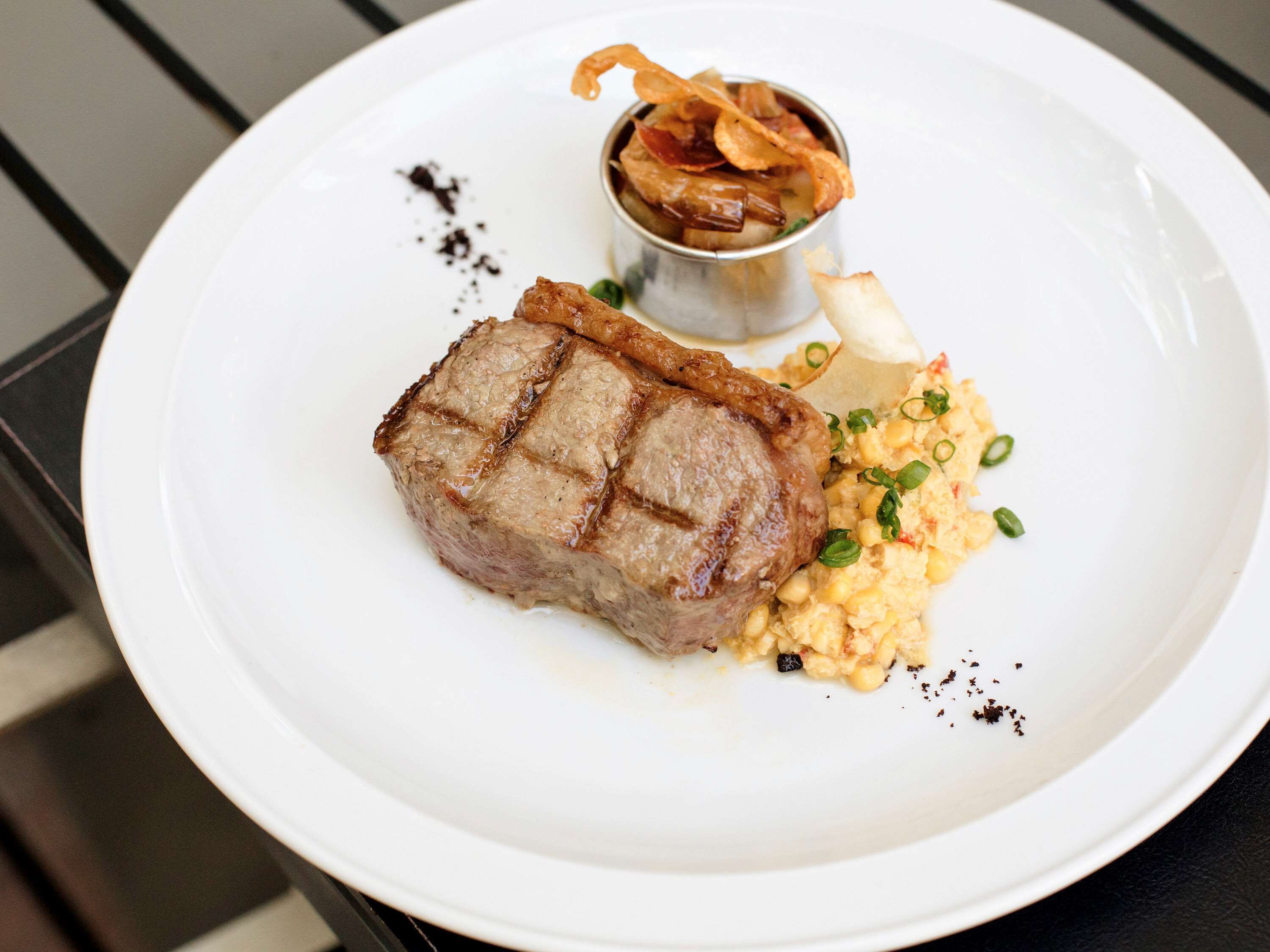 Un plato con un bistec a la parrilla, maíz y un recipiente pequeño con trocitos crujientes de tocino.