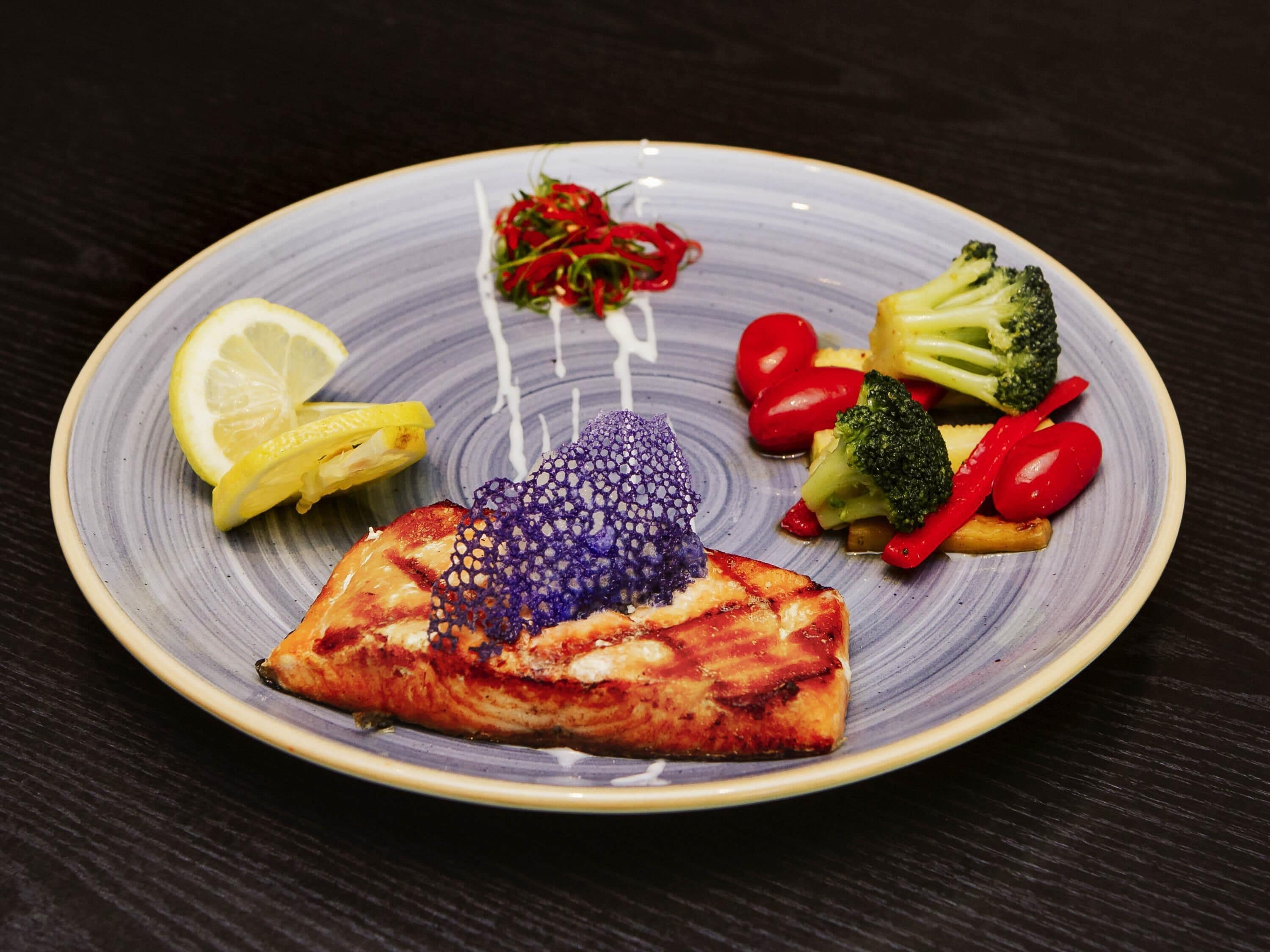 A plate with grilled salmon, lemon slices, cherry tomatoes, broccoli, and red bell peppers.