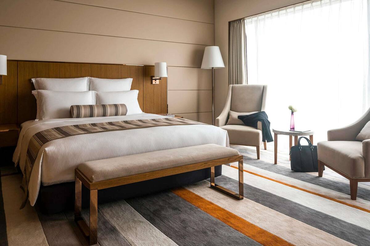 Ein Hotelzimmer mit einem großen Bett, zwei Nachttischen, einer Bank, einem Sessel, einem kleinen Tisch und einem Fenster mit Vorhängen.
