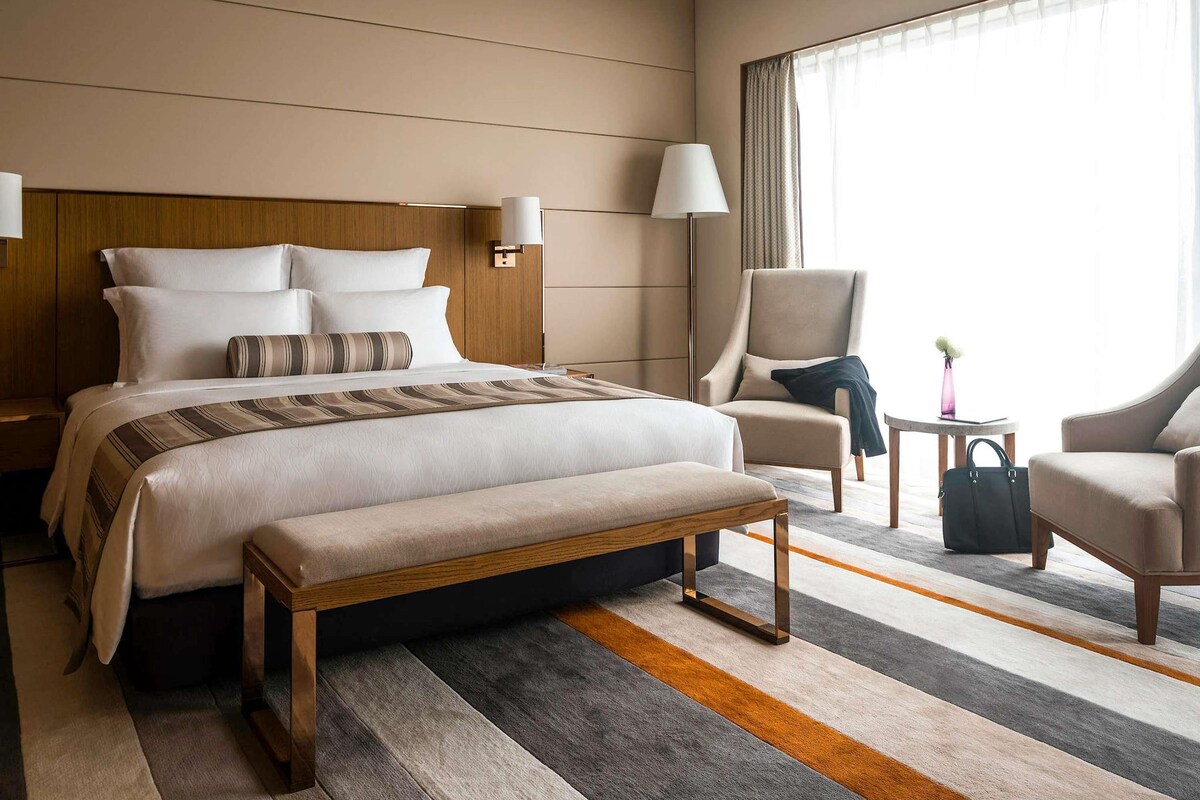 A hotel room with a large bed, two bedside tables, a bench, a chair, a small table, and a window with curtains.