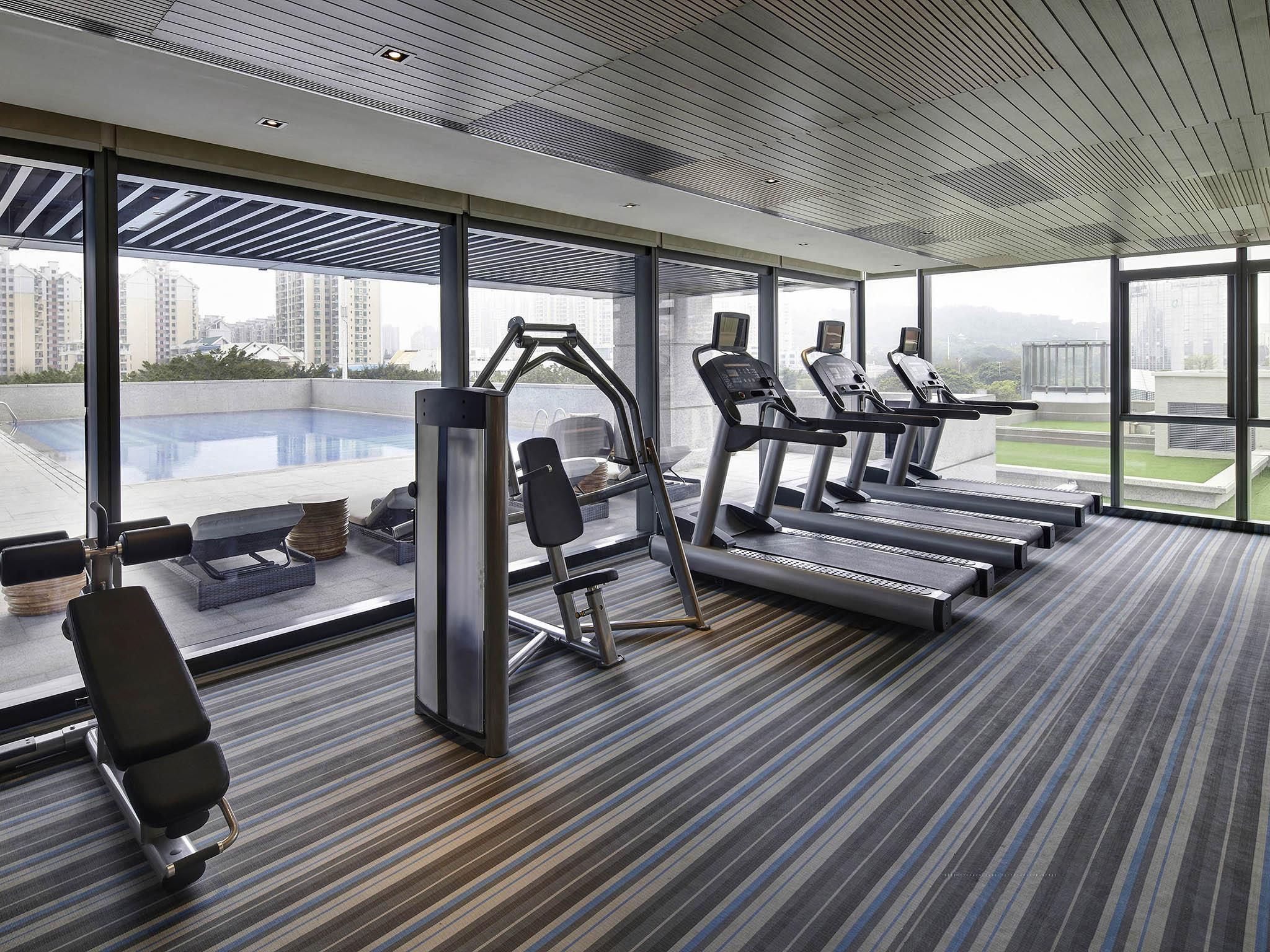 A modern gym with treadmills, a weight machine, and a bench. Large windows offer a view of the cityscape.