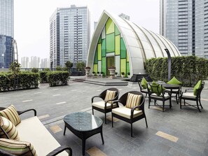 A modern outdoor seating area with wicker chairs and tables, surrounded by greenery and a distinctive green and white building. - Pullman Zhuhai (Zhuhai)