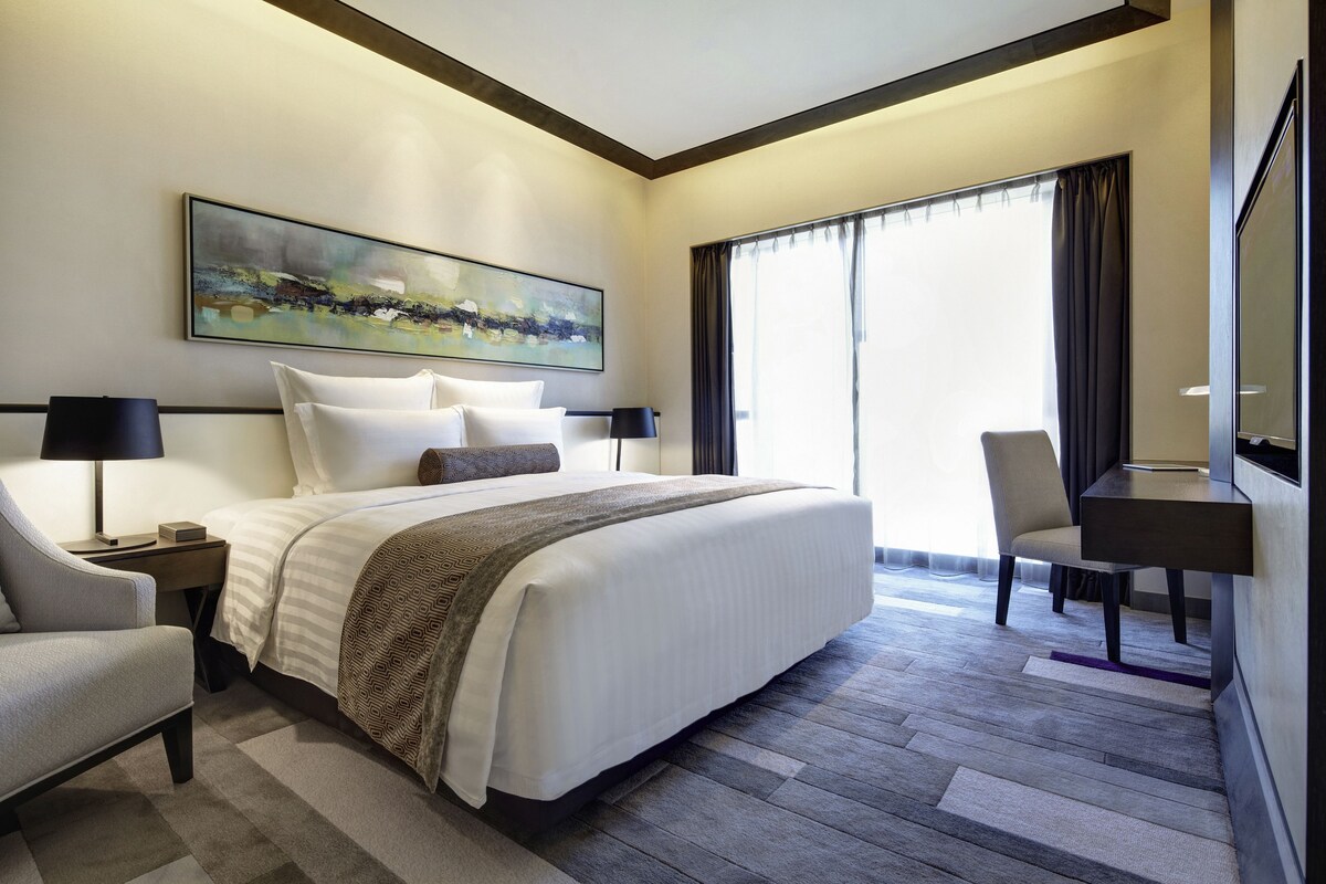 A hotel room with a large bed, a desk, a chair, and a painting on the wall.