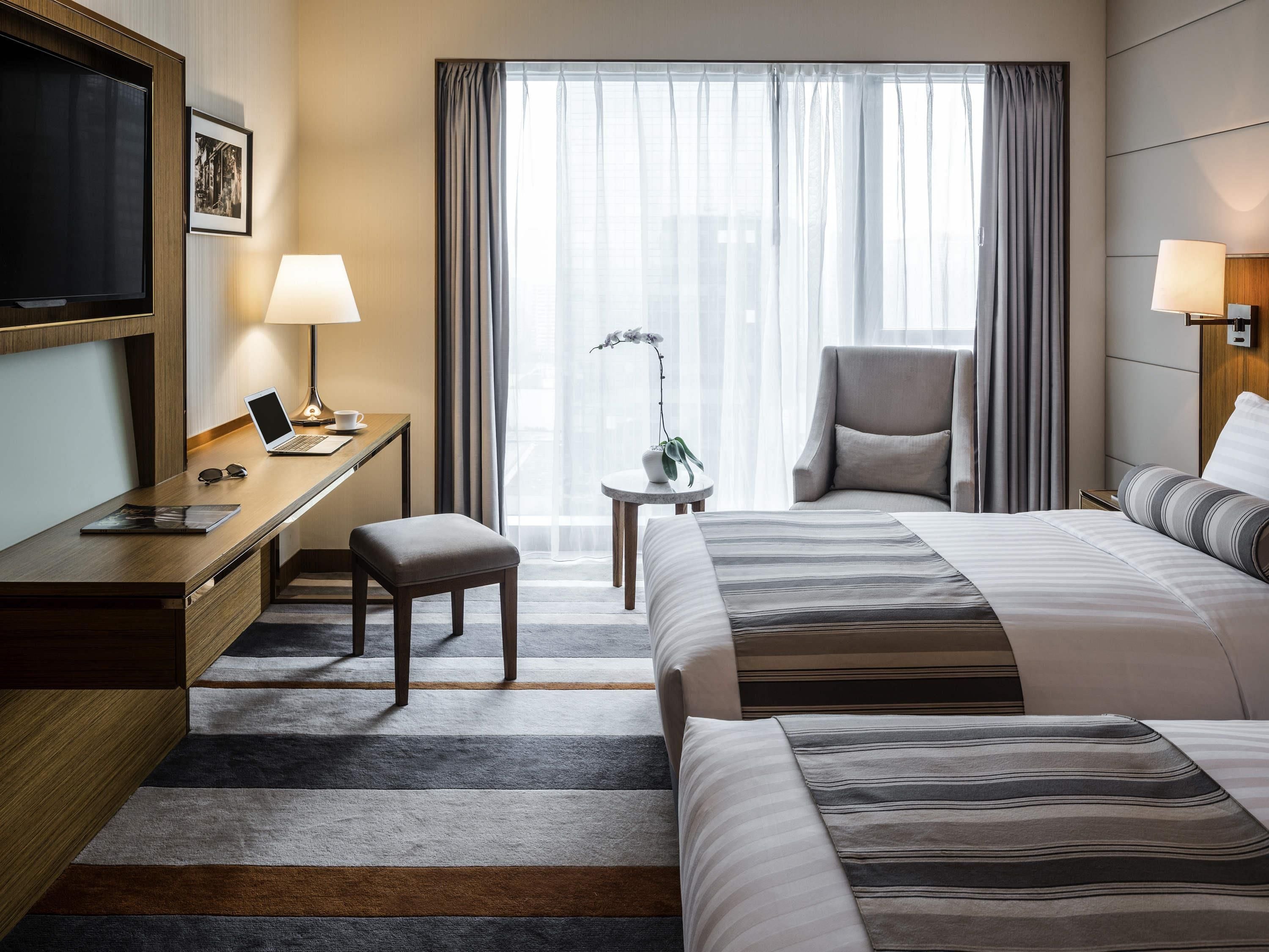 A hotel room with a bed, desk, chair, TV, lamp, and a view of the city through the window.