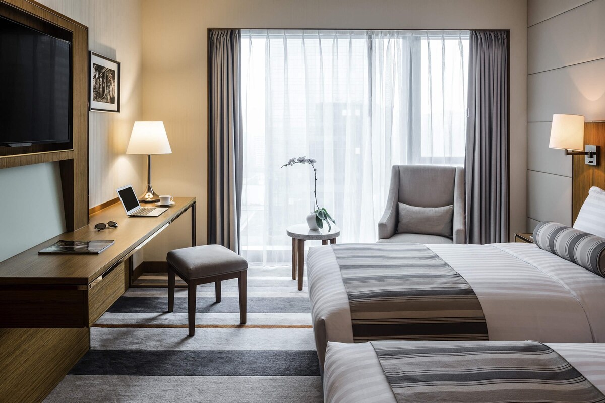 A hotel room with a bed, desk, chair, TV, lamp, and a view of the city through the window.