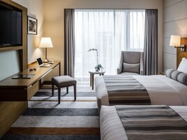 A hotel room with a bed, desk, chair, TV, lamp, and a view of the city through the window.