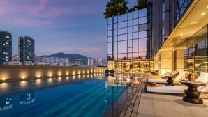 Ein Pool auf dem Dach mit freiem Blick auf die Stadtlandschaft, ein modernes Gebäude mit Glasfassade und ein gut beleuchteter Loungebereich mit Sitzgelegenheiten und Bar.