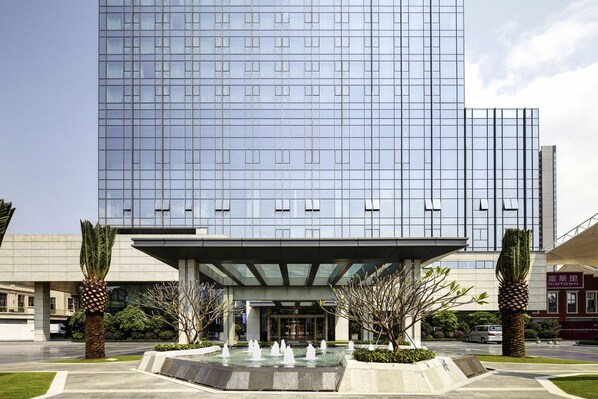 A modern building with a glass facade, a covered entrance, and a water feature in front. - Pullman Zhuhai (Zhuhai)