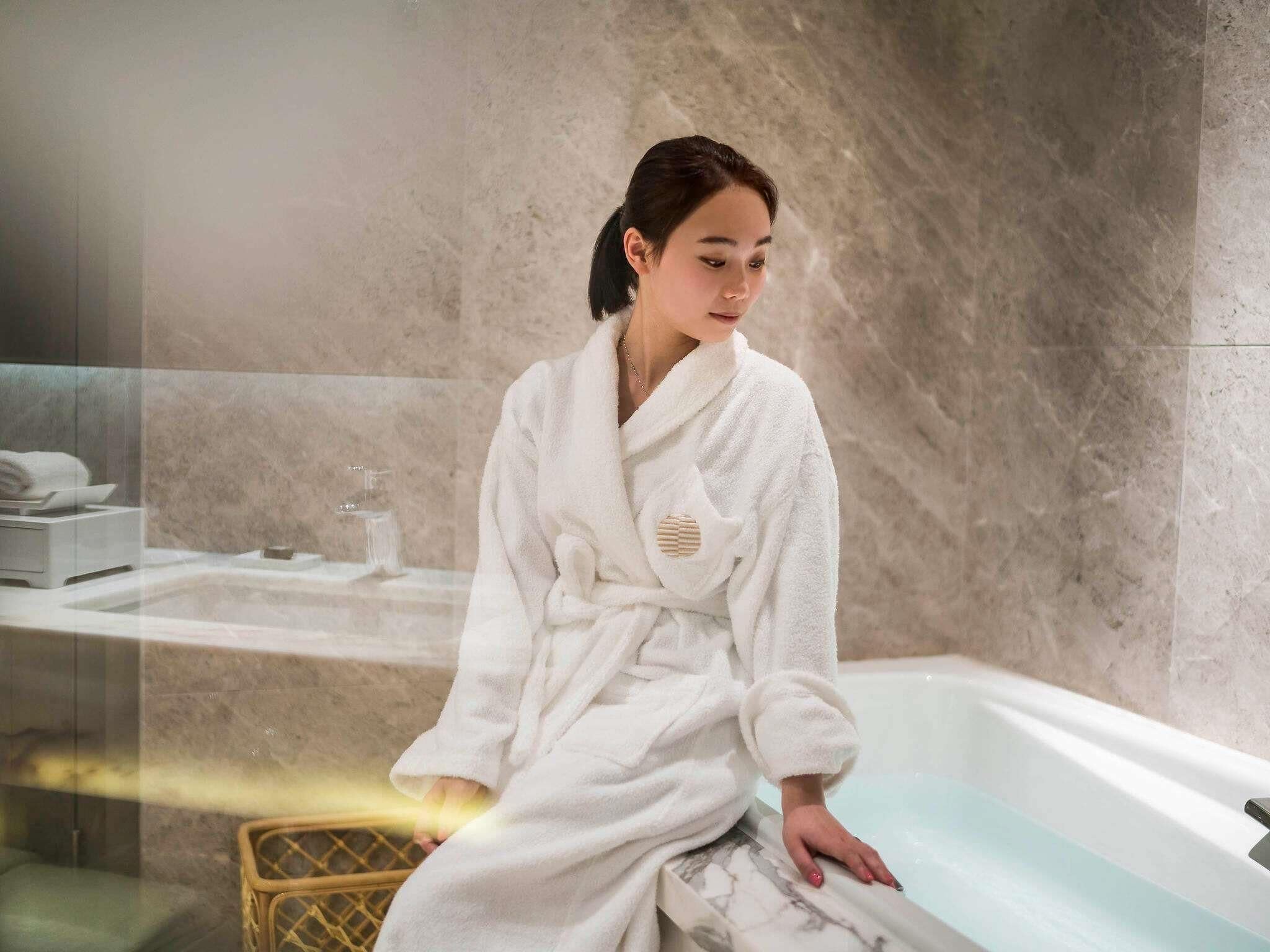 A person in a white bathrobe sitting by a bathtub in a bathroom with marble walls and a glass partition.