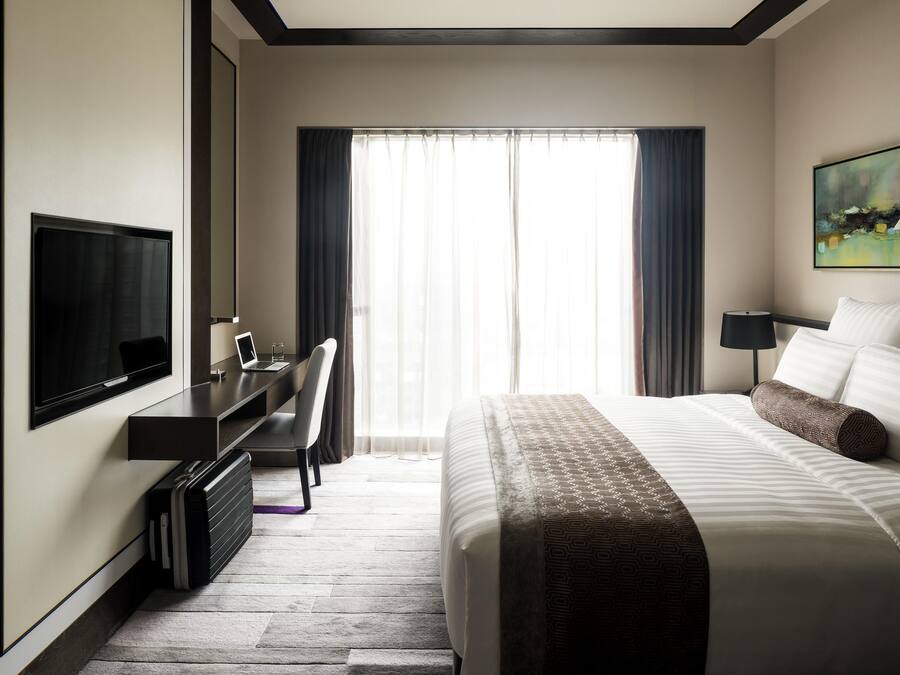 A hotel room with a bed, a desk with a laptop, a TV, and a large window with curtains.