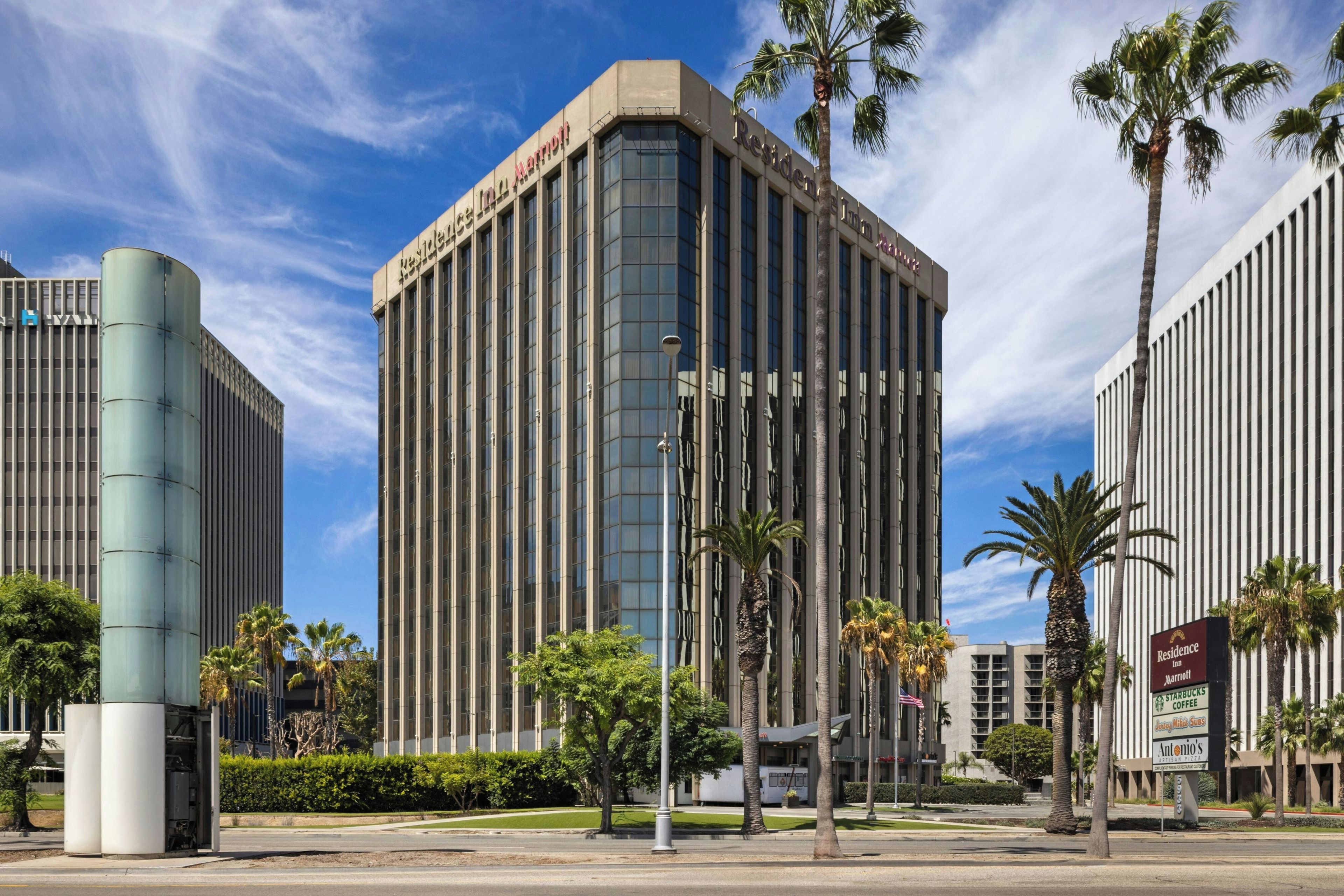 A modern hotel building with a prominent 'Residence Inn' sign, surrounded by palm trees and other buildings.
