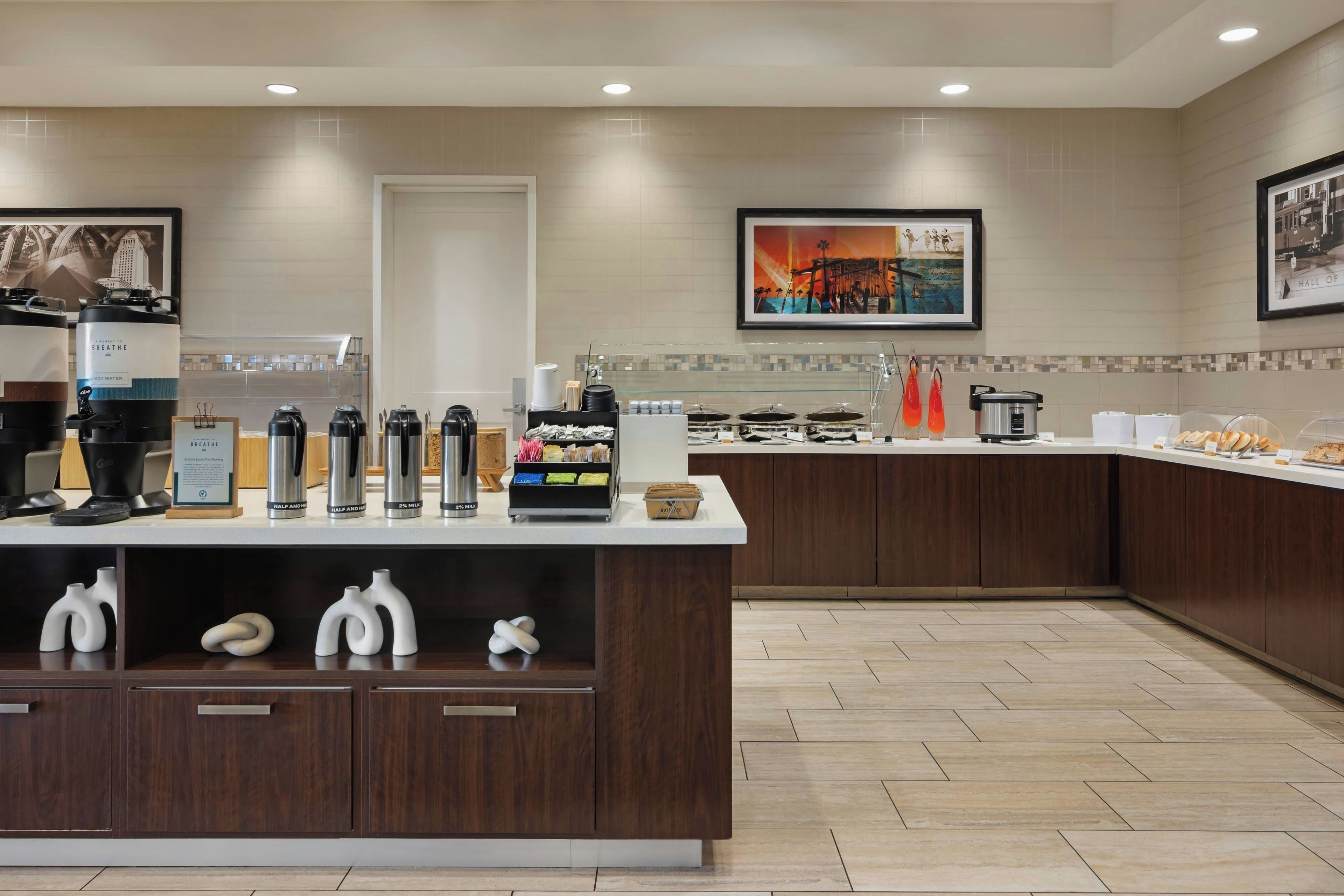 A modern hotel breakfast area with a coffee station, a variety of food options, and artwork on the walls.