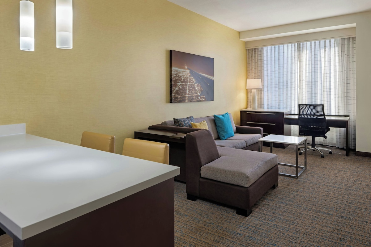 Una habitación de hotel con escritorio, sofá, sillón, una mesita y ventana con cortinas.