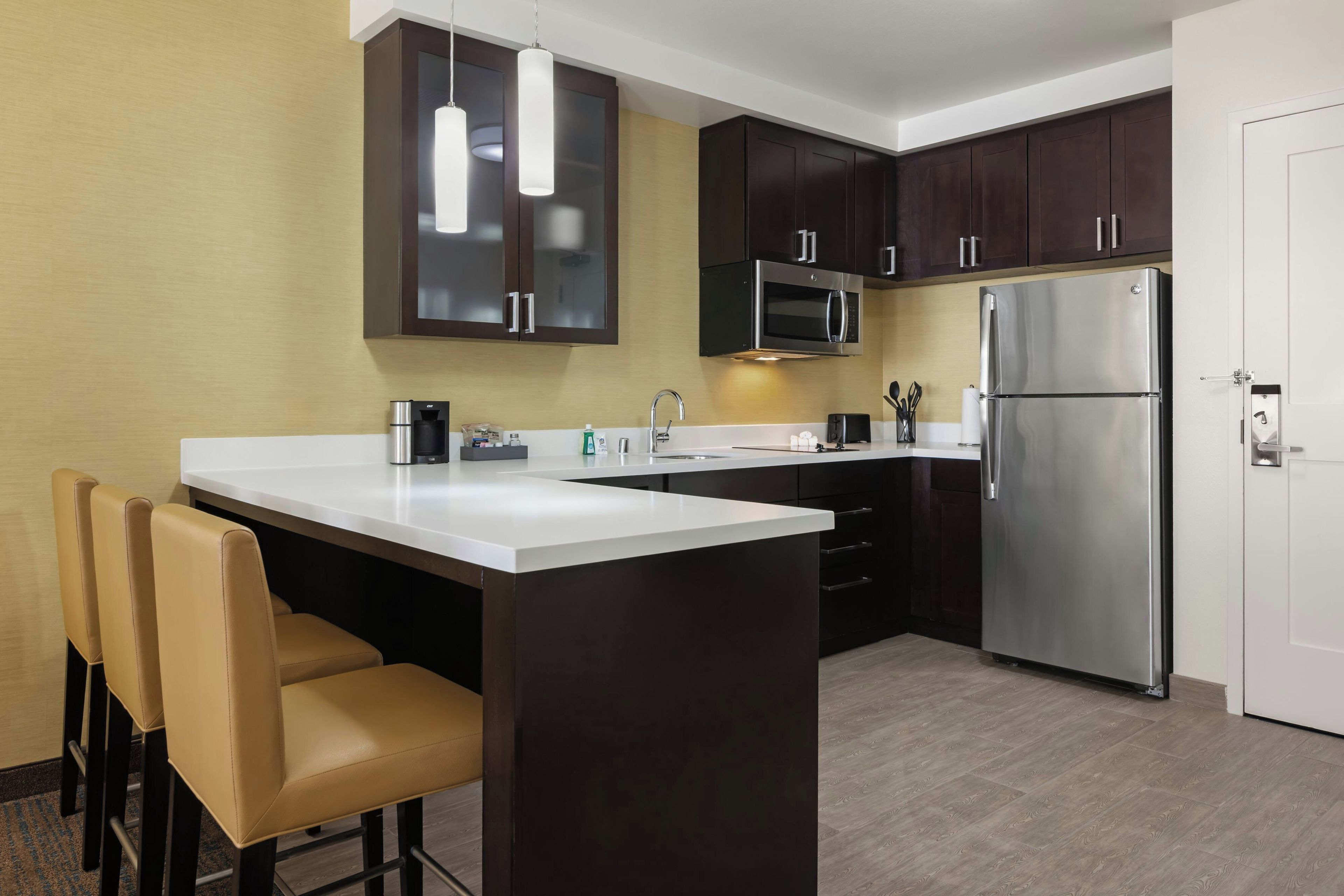 A modern kitchen with dark cabinets, stainless steel appliances, a white countertop, and a seating area with two chairs.