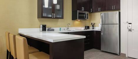 A modern kitchen with dark cabinets, stainless steel appliances, a white countertop, and a seating area with two chairs.