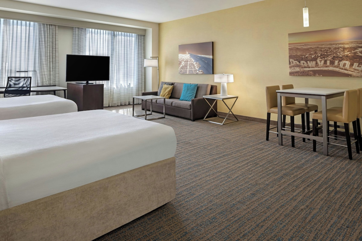 Habitación de hotel con dos camas, televisión, escritorio con silla, sofá y lámpara.