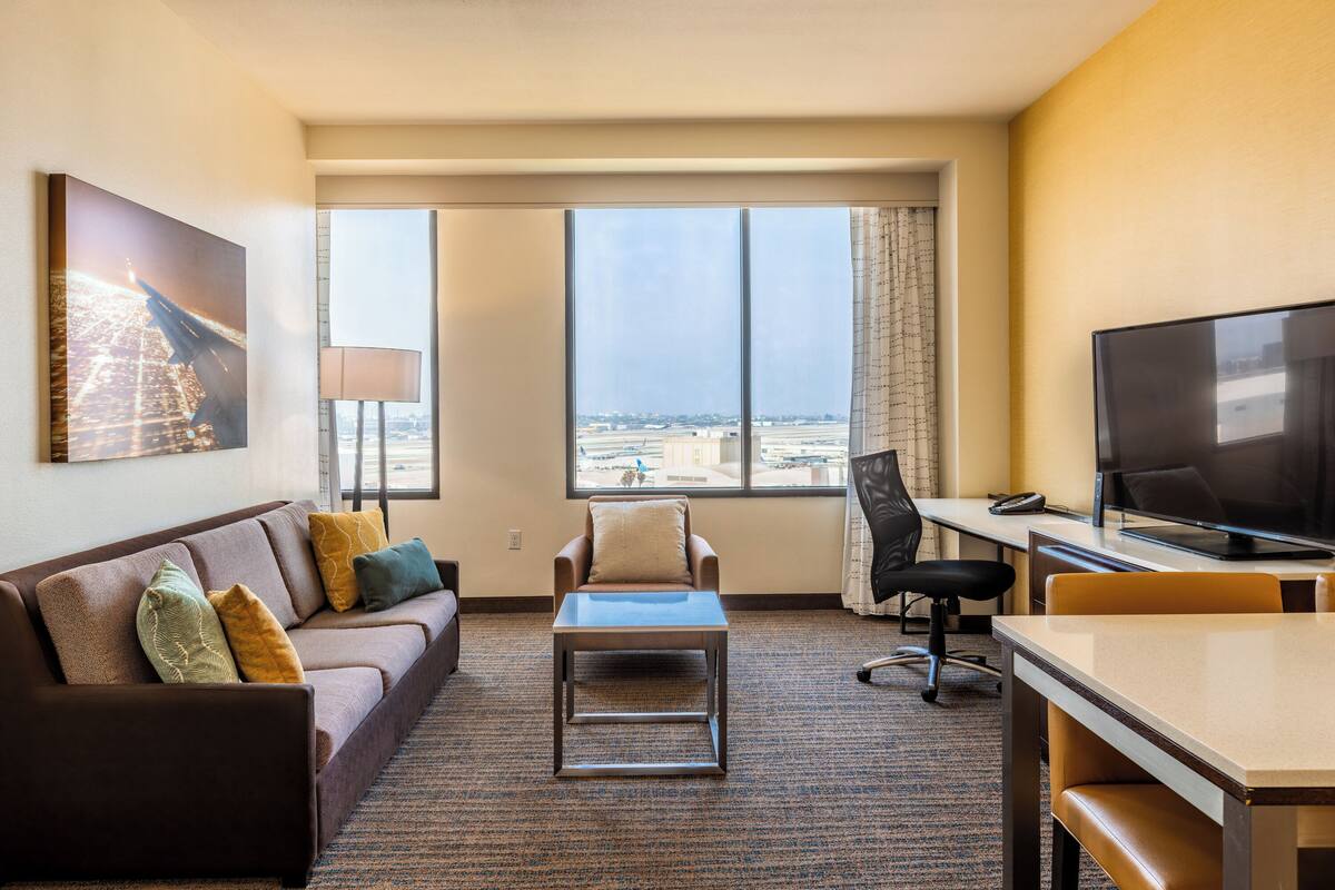 A hotel room with a sofa, a small table, a TV, and a large window with a city view.