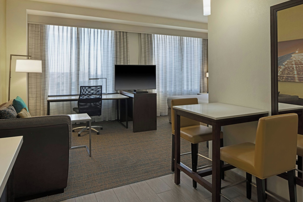 Habitación de hotel con escritorio, silla, sofá, televisor y ventana con cortinas.