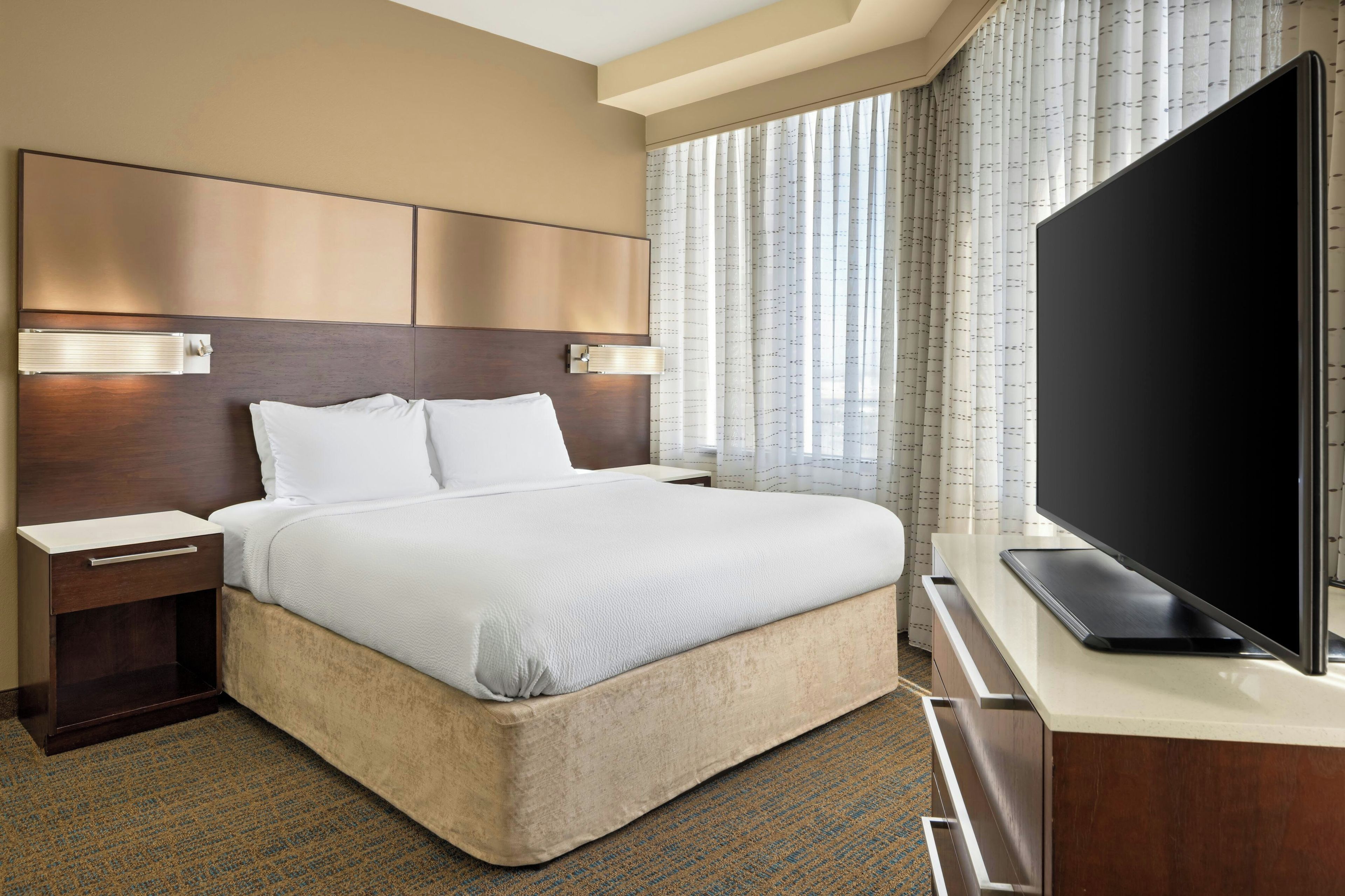 A hotel room with a bed, bedside tables, a flat-screen TV, and a large window with curtains.