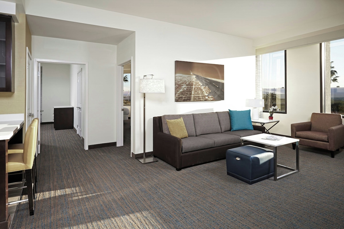 Una habitación de hotel moderna con sofá, sillón y una pequeña mesa. Hay un cuadro en la pared y una ventana con vistas.