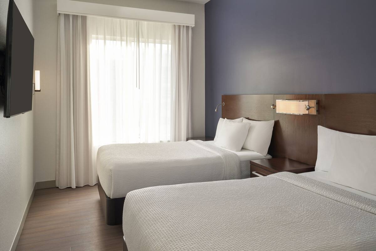 A hotel room with two beds, a TV mounted on the wall, and a window with sheer curtains.