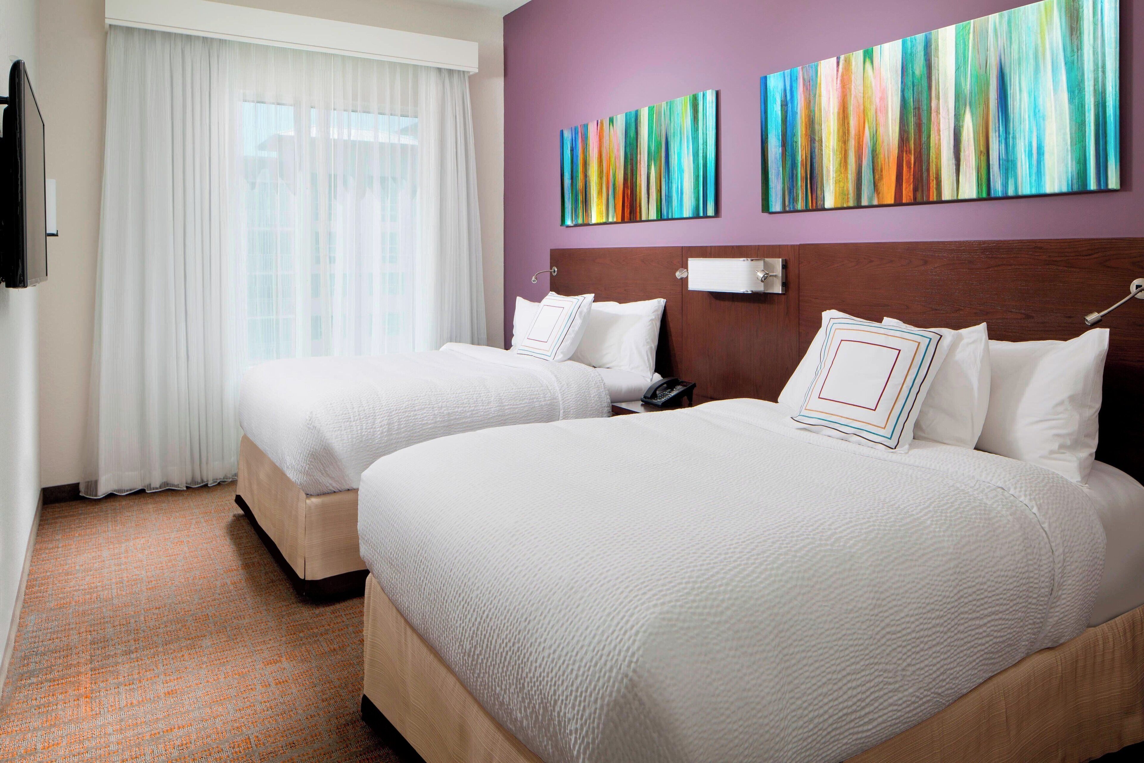 A hotel room with two beds, a large window with white curtains, and two colorful abstract paintings on the wall.