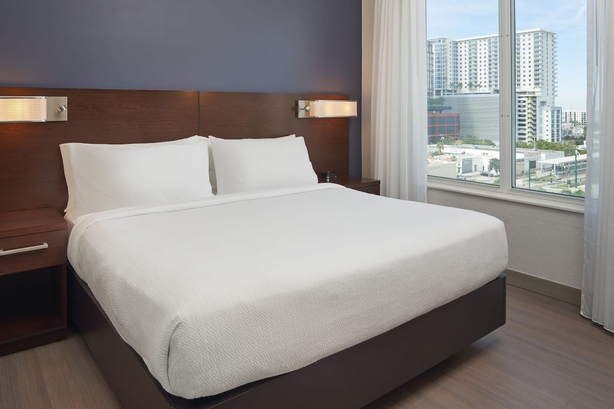 A hotel room with a large bed, wooden headboard, bedside lamps, and a view of city buildings through a window.