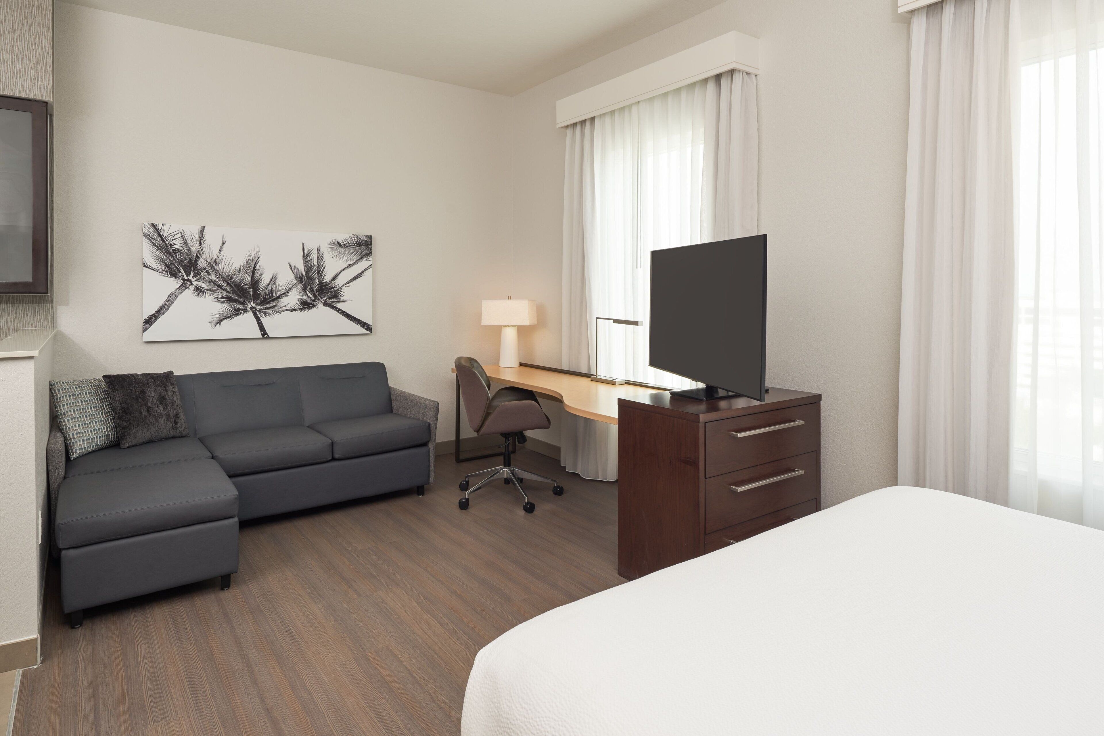 Habitación de hotel con cama, escritorio con silla, sofá, televisor y un cuadro en la pared.