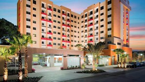 A multi-story hotel with a modern design, featuring balconies with red railings, a parking area, and palm trees. - Residence Inn by Marriott West Palm Beach Downtown (West Palm Beach)
