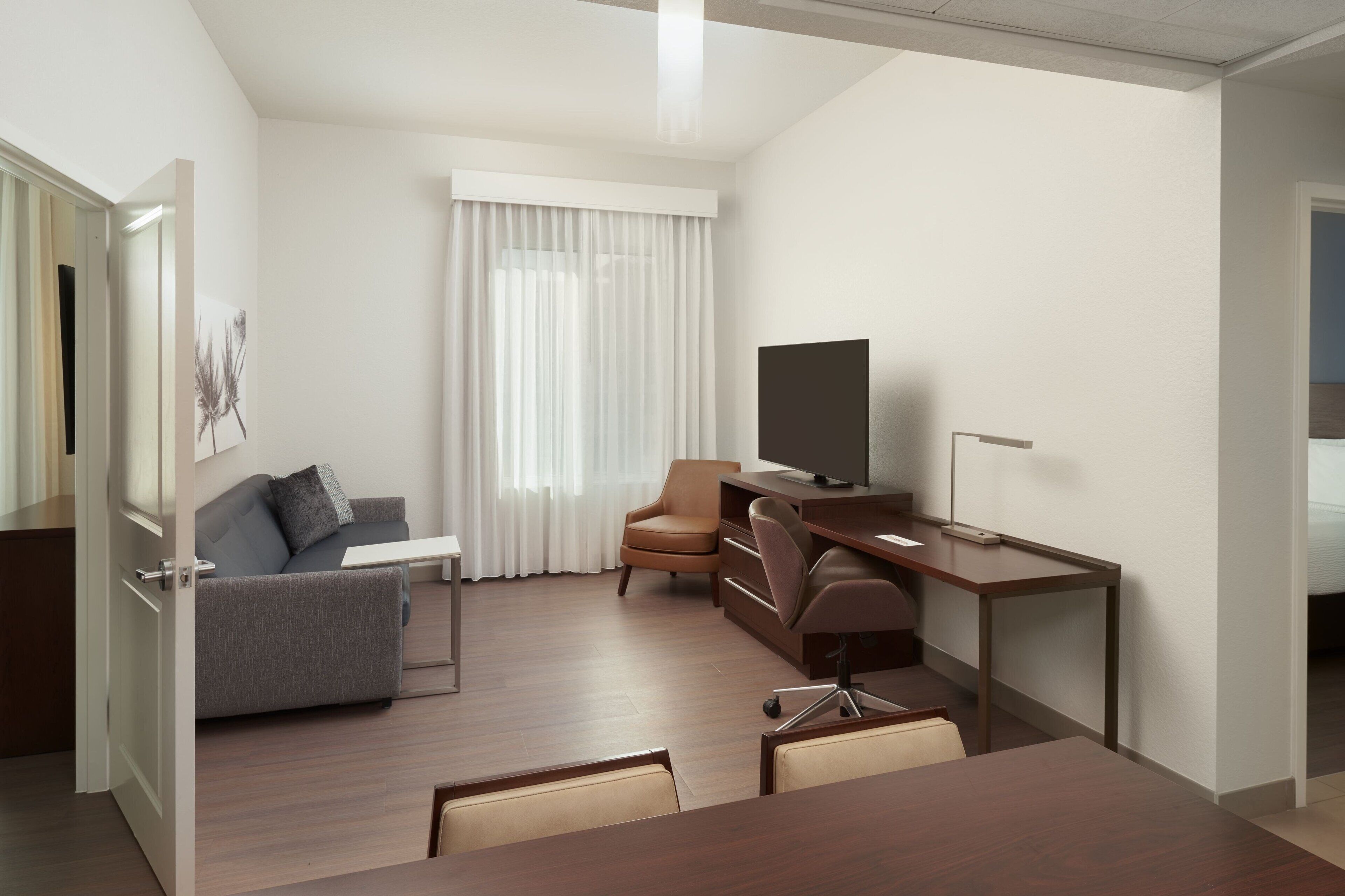 A modern hotel room with a sofa, a desk, a television, and a large window with curtains.