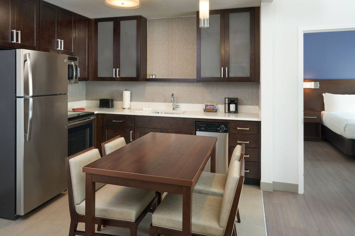 A modern kitchen with a stainless steel refrigerator, a built-in oven, and a dishwasher. There is a wooden dining table with four chairs.
