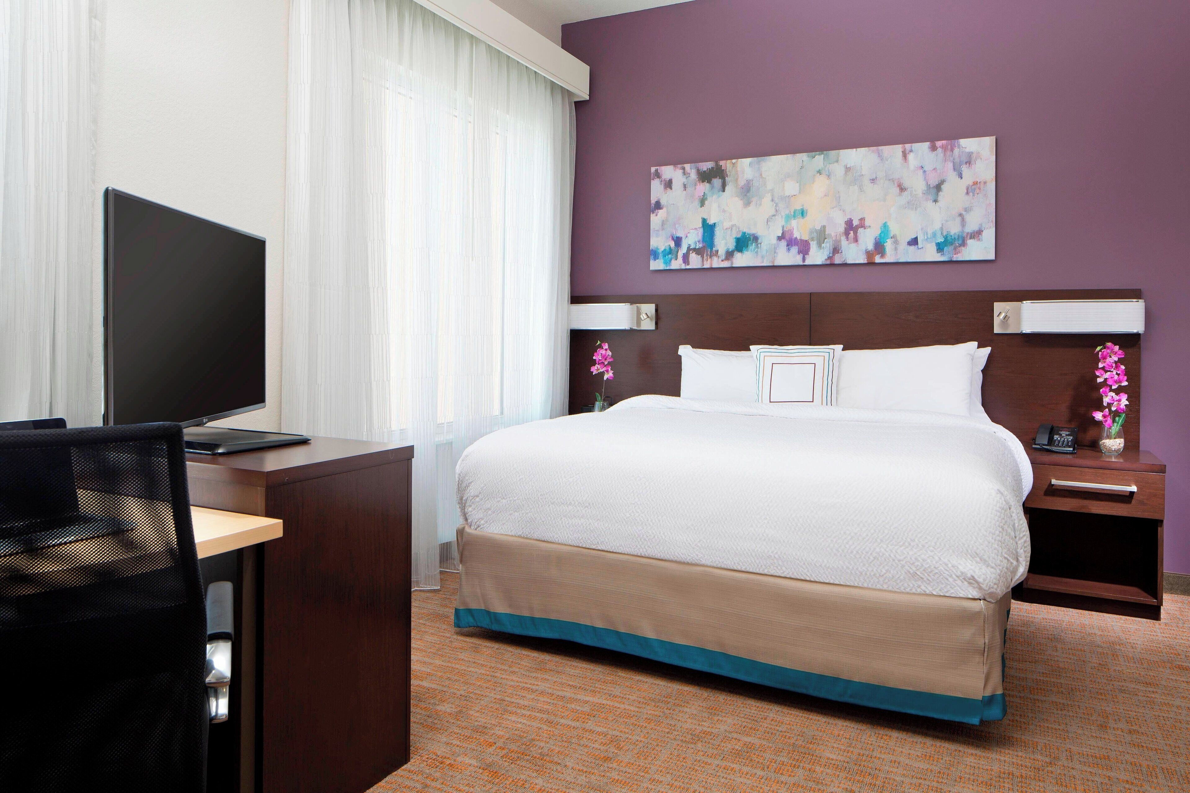 A hotel room with a bed, a desk with a computer, a TV, and a nightstand with a phone and flowers.