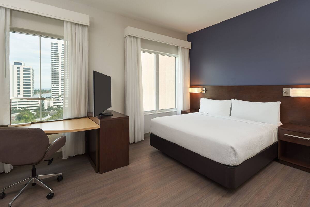 A hotel room with a large bed, a desk with a chair, a TV, and a view of tall buildings.