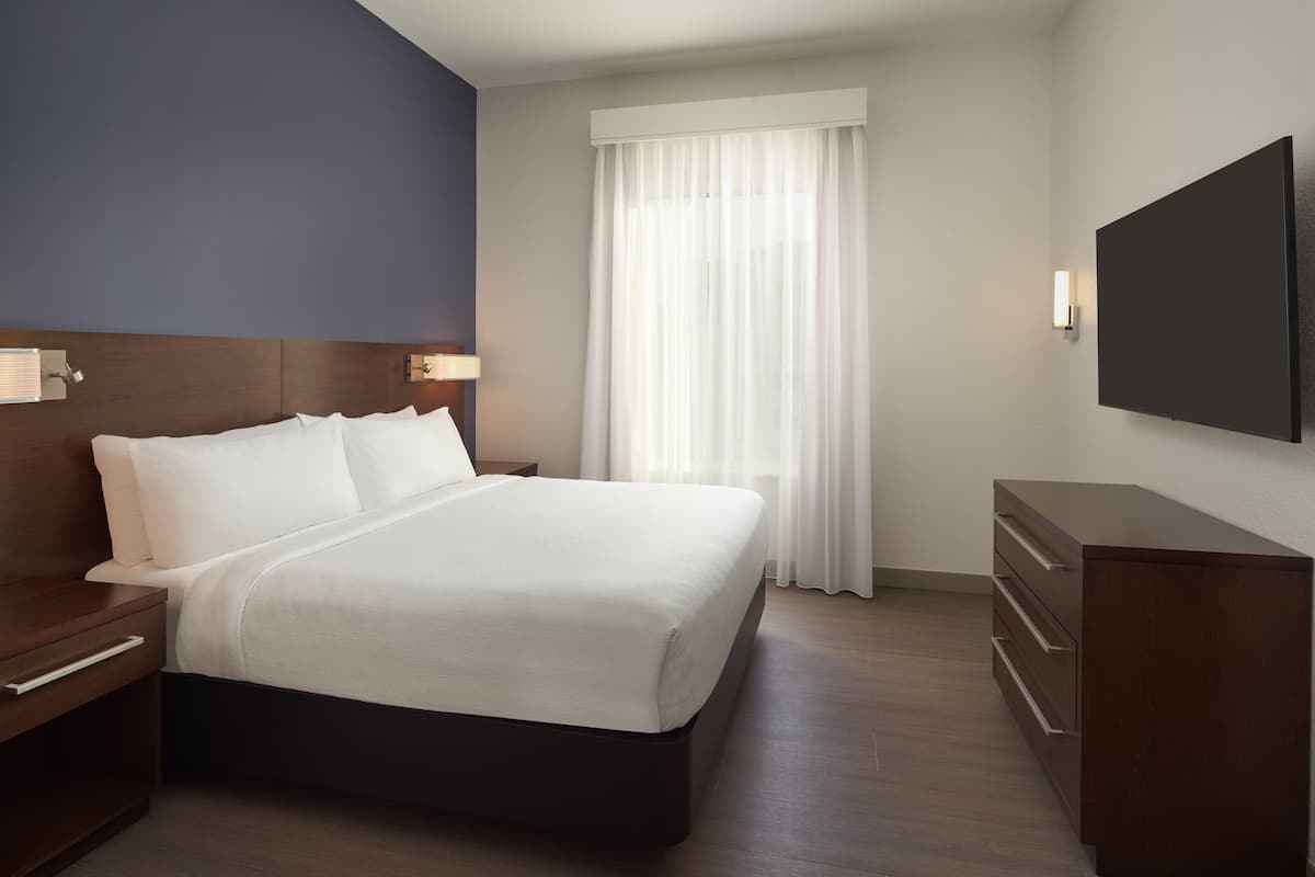 A modern hotel room with a large bed, wooden headboard, a flat-screen TV, a nightstand, and a dresser.