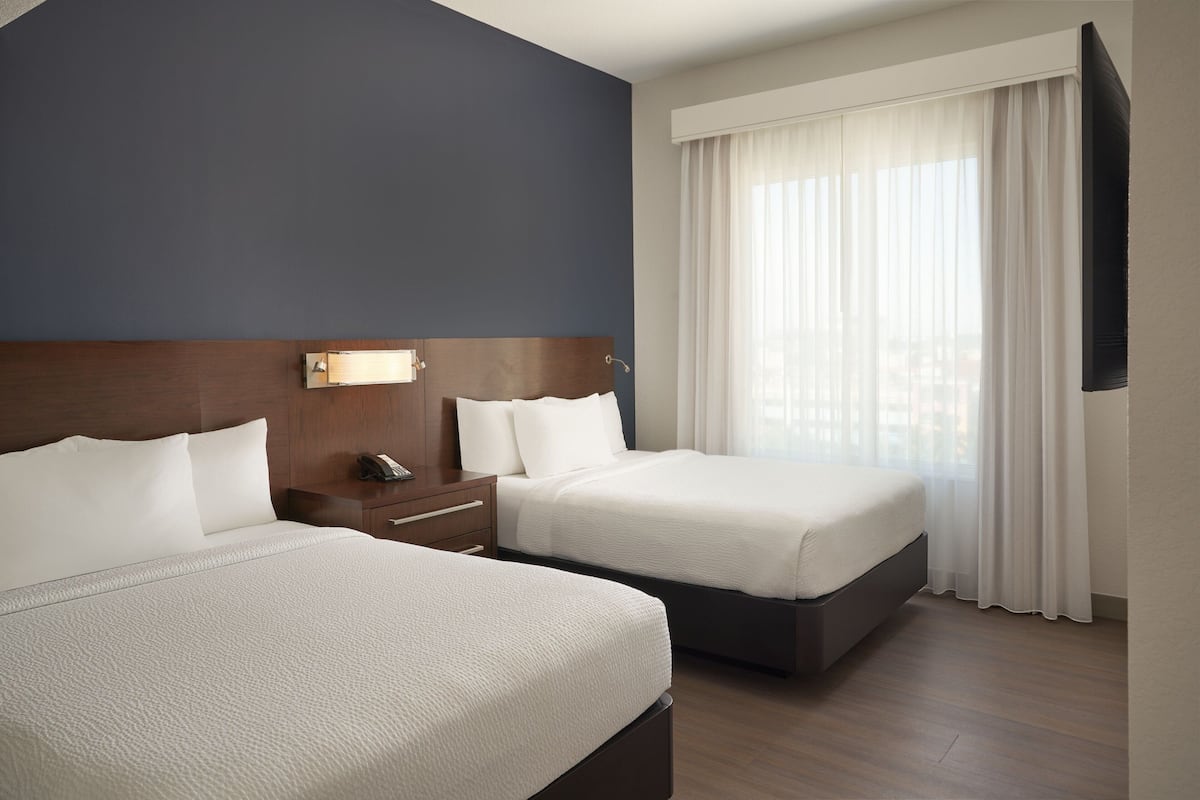A hotel room with two beds, a wooden headboard, a bedside table with a lamp, and a large window with sheer curtains.