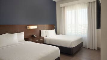 A hotel room with two beds, a wooden headboard, a bedside table with a lamp, and a large window with sheer curtains.