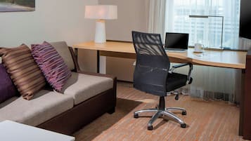 Una habitación de hotel con sofá, escritorio, laptop, silla, lámpara y televisor.