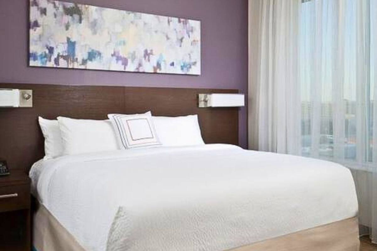 A hotel room with a large bed, a headboard with a light fixture, a nightstand, and a window with sheer curtains.