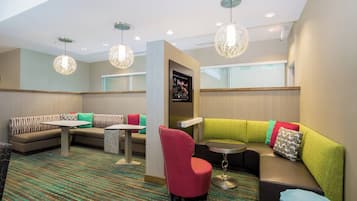 Um hall de hotel com um sofá em canto, uma poltrona vermelha, uma mesinha e luminárias pendentes.