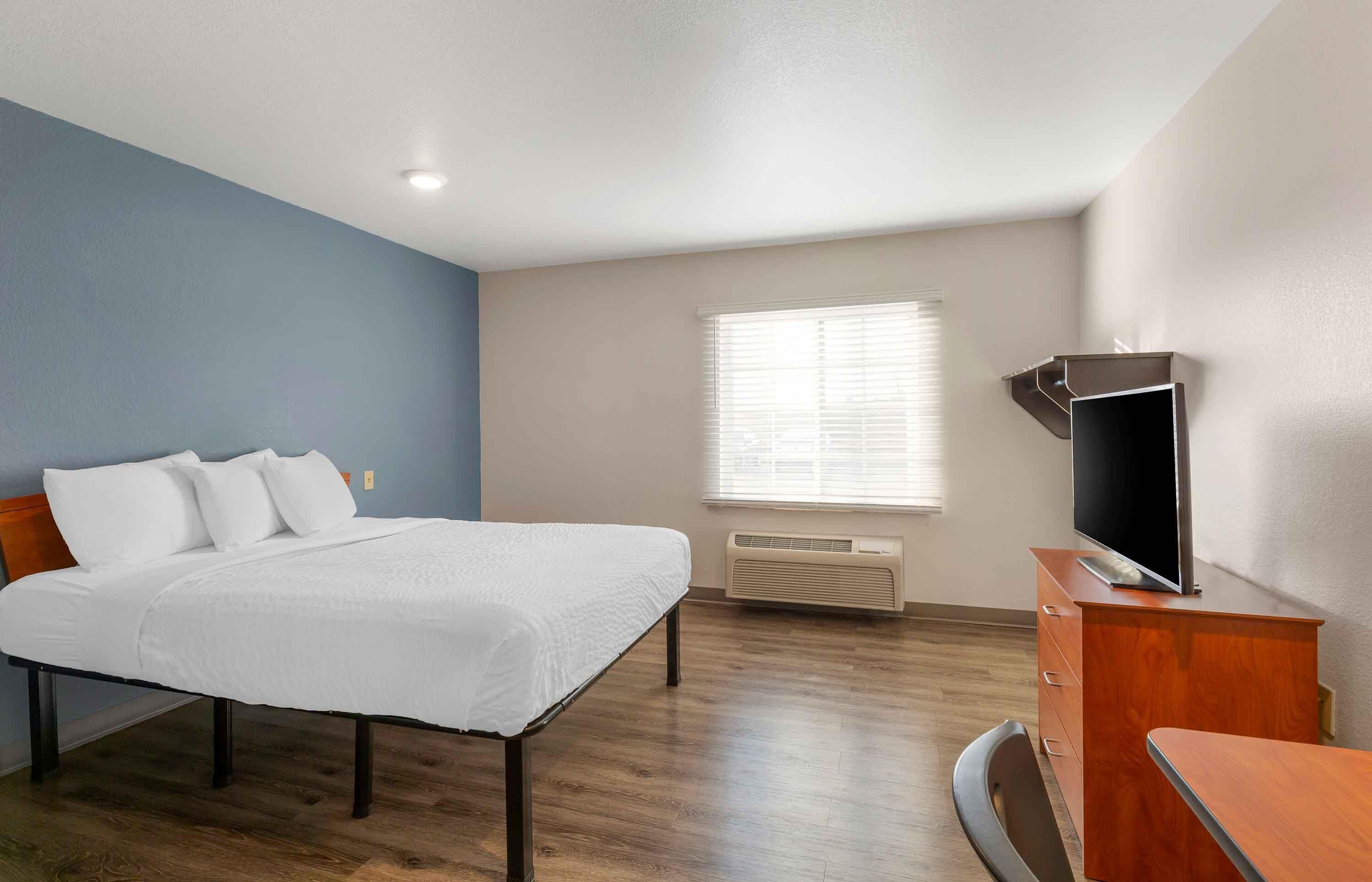 A hotel room with a bed, a TV, a desk, and a window with blinds.
