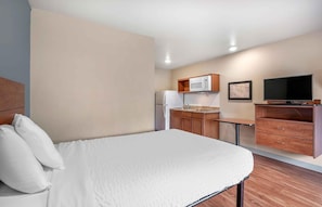 A compact hotel room with a bed, a kitchenette, a flat-screen TV, and a small seating area.
