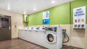 A laundry facility with multiple washing machines, a wall-mounted soap and bleach dispenser, and a sign advertising a mobile app for laundry services. - Extended Stay America Select Suites - Dayton - Miamisburg (Dayton)