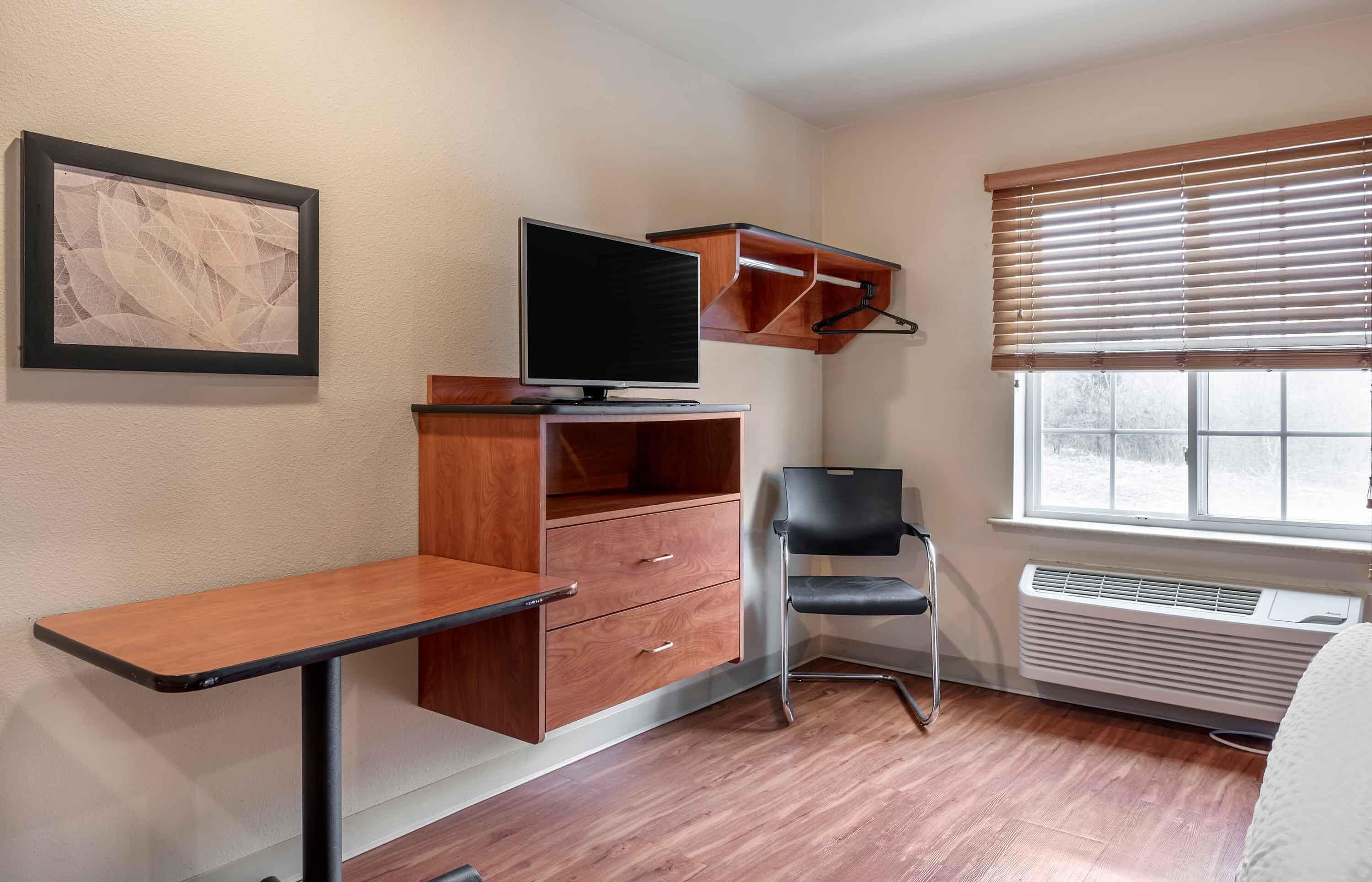 A compact room with a desk, a chair, a television, a heater, and a window with blinds.
