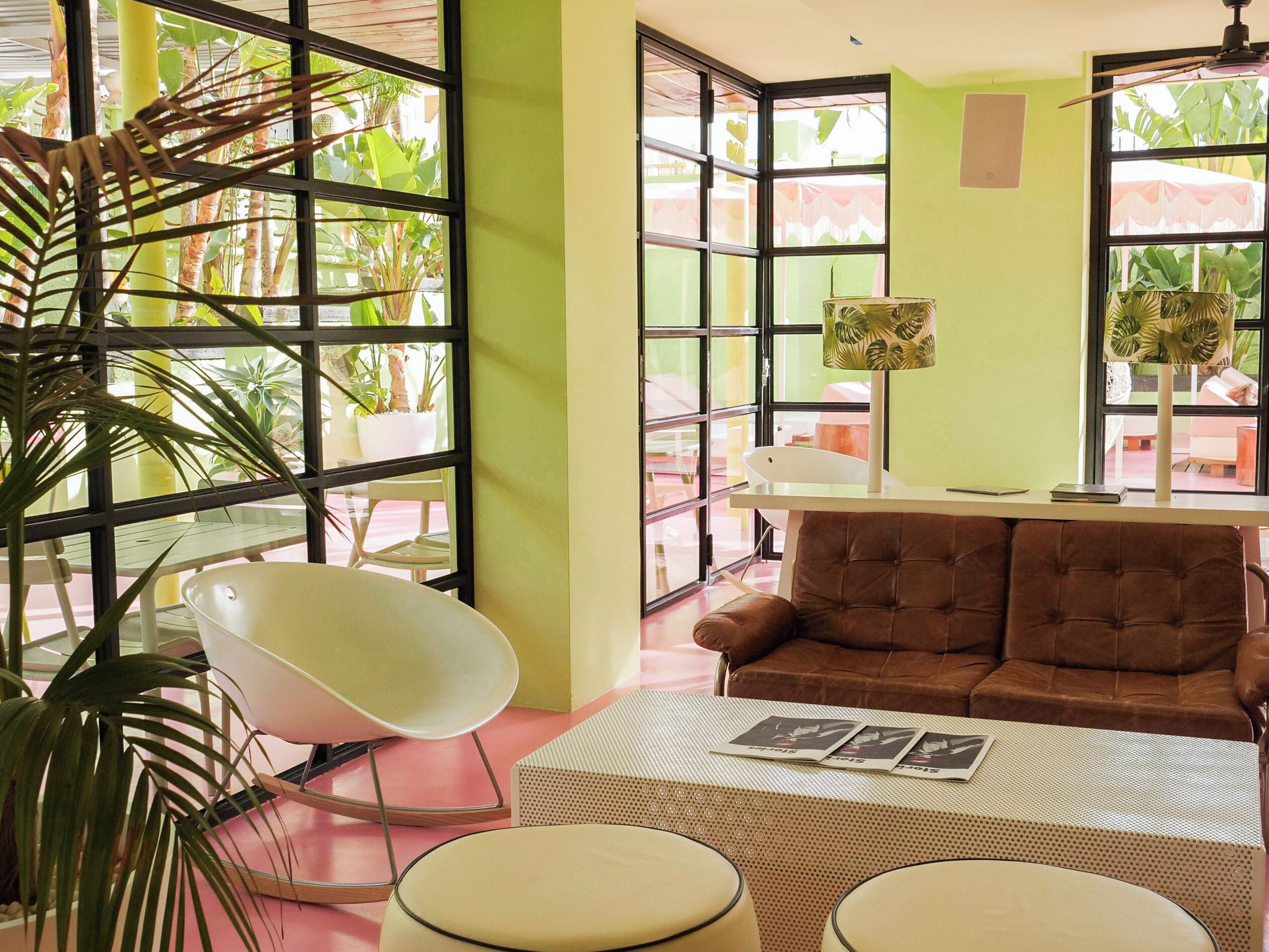 Una habitación con un sofá marrón, una silla blanca, una mesa con revistas y ventanas amplias con plantas afuera.