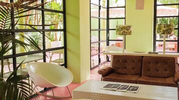 Una habitación con un sofá marrón, una silla blanca, una mesa con revistas y ventanas amplias con plantas afuera.