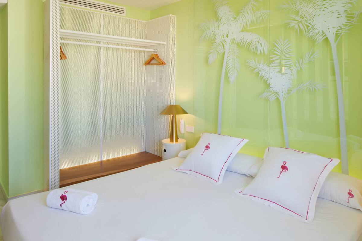 Quarto de hotel com cama, mesa de cabeceira, luminária e painel decorativo com silhuetas de palmeiras.