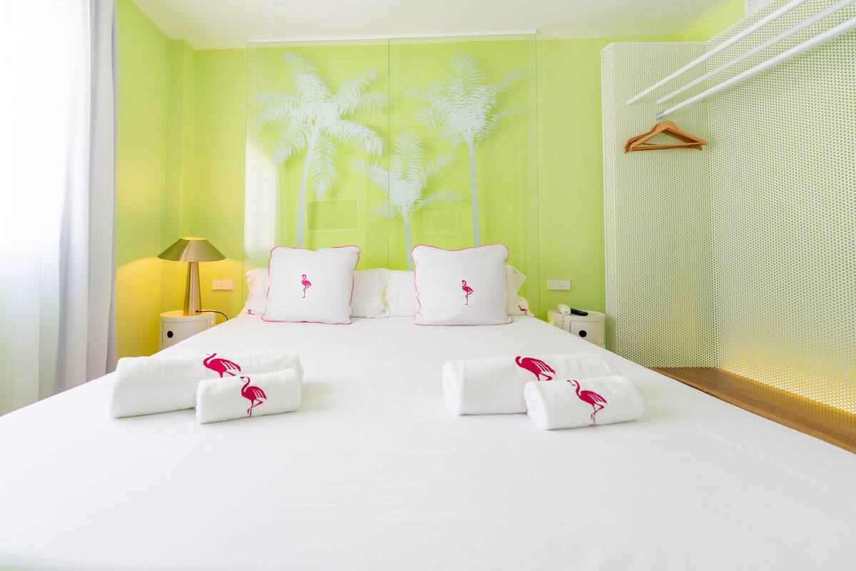 Quarto de hotel com cama, almofadas brancas, mesa de cabeceira, luminária e parede com desenho de palmeira.